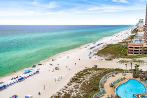 A beach resort with a swimming pool, beach chairs, umbrellas, and a pier in the background.