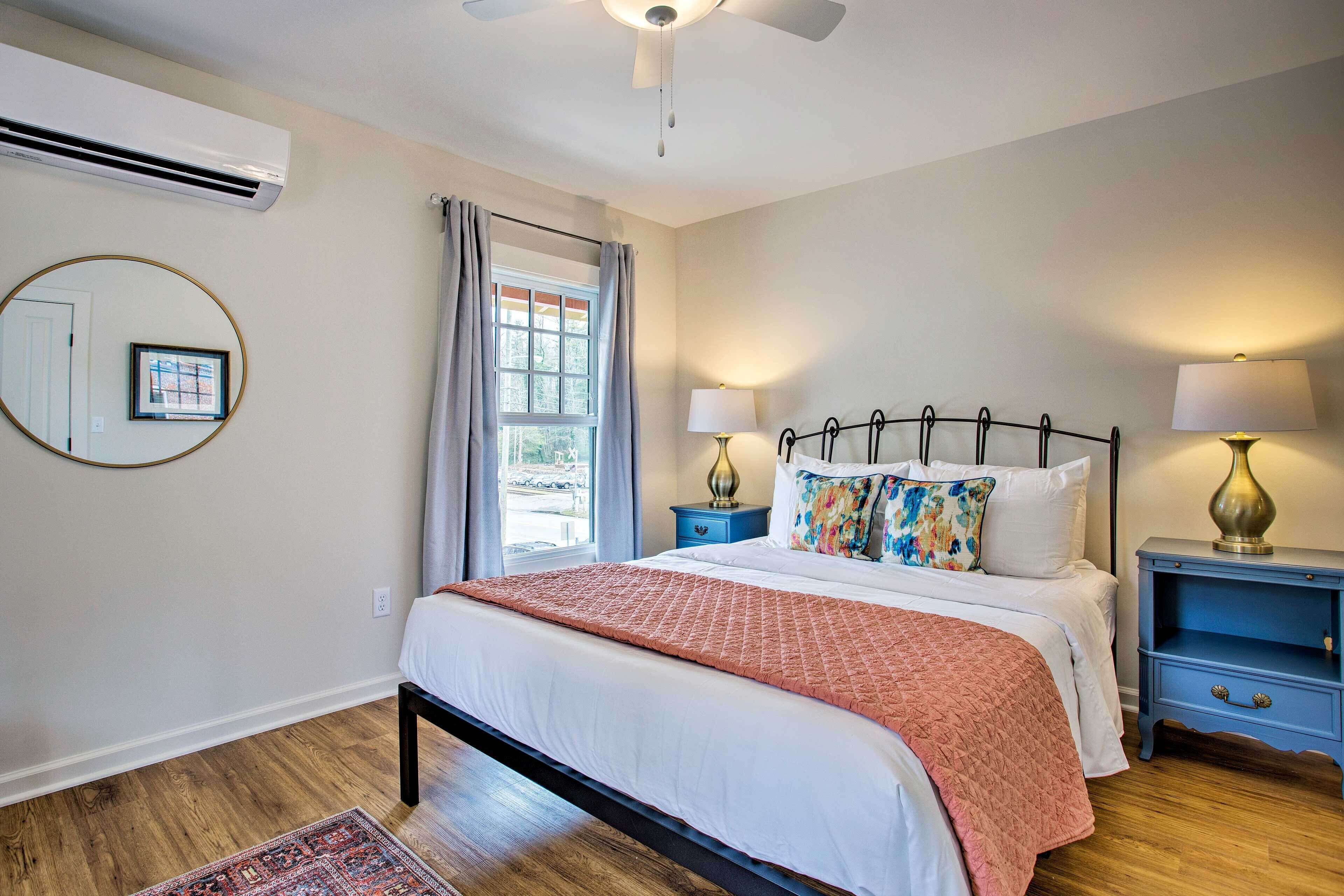 A bedroom with a bed, two bedside tables, a nightstand, a window with curtains, an air conditioning unit, and a wall-mounted mirror.