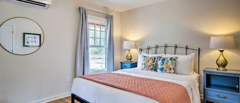 A bedroom with a bed, two bedside tables, a nightstand, a window with curtains, an air conditioning unit, and a wall-mounted mirror.