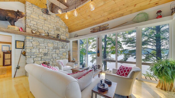 A cozy living room with a stone fireplace, wooden ceiling, and large windows overlooking a lake. - Beautiful Embden Pond Cabin w/ Dock & Kayaks! (North Anson)