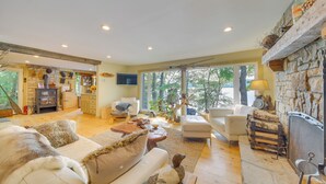 A spacious living room with a fireplace, a stone wall, and a large window overlooking the outdoors. - Beautiful Embden Pond Cabin w/ Dock & Kayaks! (North Anson)