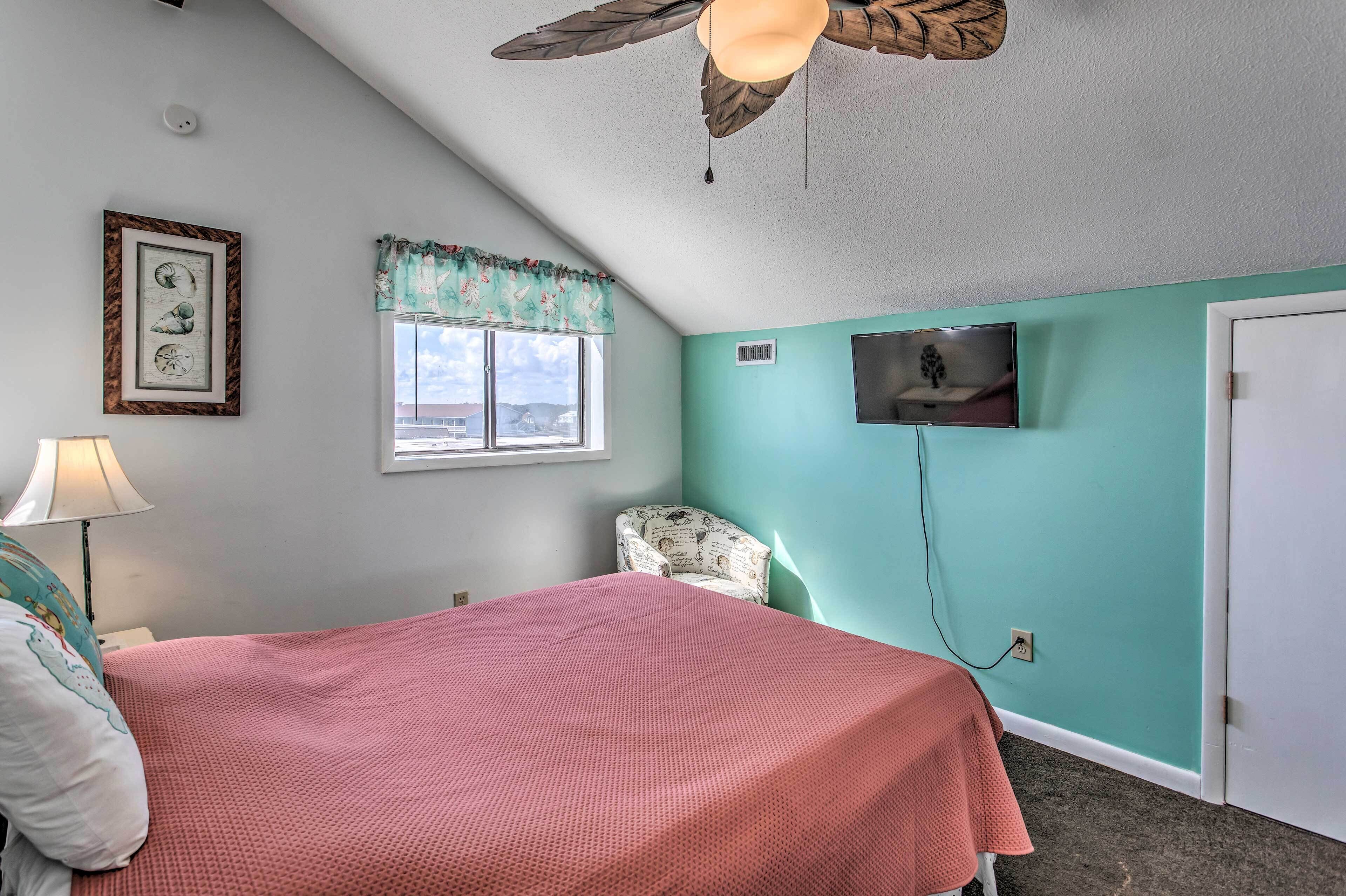Une chambre avec un lit, un ventilateur de plafond, une fenêtre avec un rideau à motifs floraux, un téléviseur fixé au mur et une porte.
