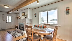 A cozy dining area with a brick fireplace, wooden chairs, and a large window offering a view of the outdoors. - Cozy Postville Retreat w/ Fireplaces & Yard! (Postville)