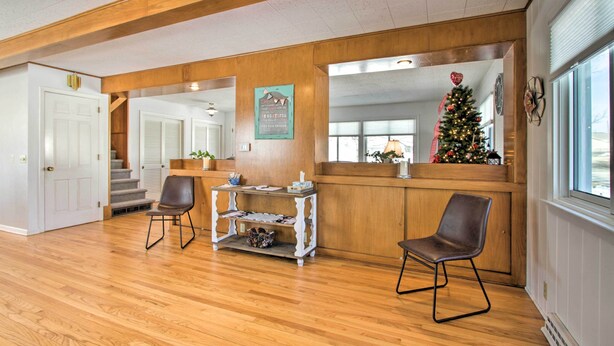 A spacious room with wooden floors, a wooden reception desk, two chairs, a small table, and a Christmas tree. - Cozy Postville Retreat w/ Fireplaces & Yard! (Postville)
