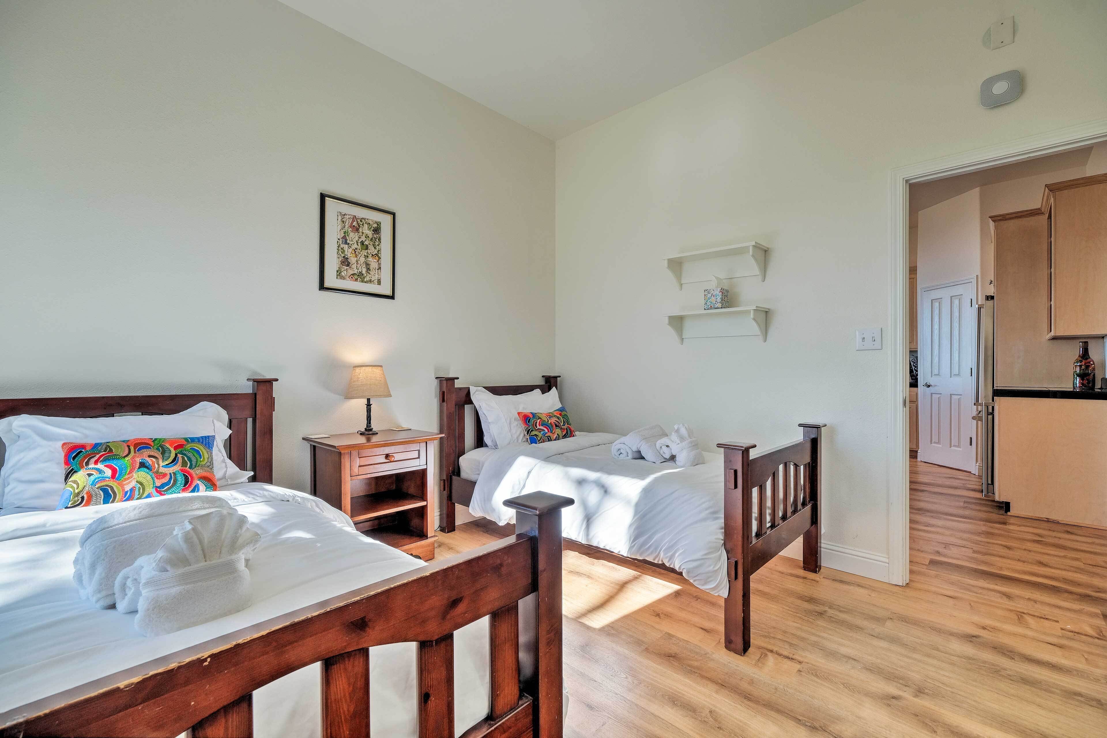 A hotel room with two beds, wooden headboards, a bedside table with a lamp, a framed picture on the wall, and a kitchen area visible through an open door.
