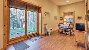 A dining area with a wooden table and chairs, a television on a stand, and a view of the outdoors through large sliding glass doors.