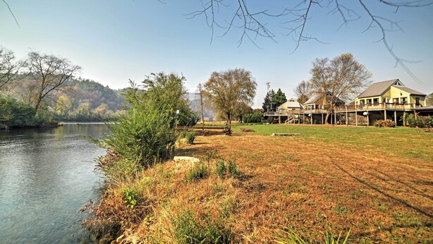 Una propiedad junto al río con un césped, una terraza de madera y un edificio con un amplio ventanal con vistas al agua.