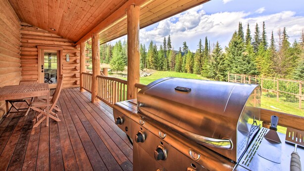 A wooden cabin with a covered porch, a grill, and a view of a forest. - Waterfront Log Home w/ 95 Acres on Yaak River (Troy)