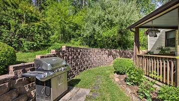 Un muro de piedra, una parrilla y una terraza de madera con una casa al fondo.
