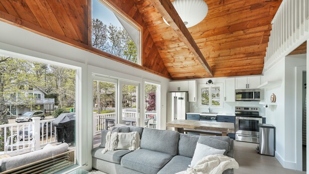 A spacious living area with a grey sofa, a glass coffee table, and a wooden ceiling. A kitchen with white cabinets and stainless steel appliances is visible in the background. - Family Friendly 5 Bedroom, Walk to Town, Beach & Ferry (Tisbury)