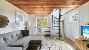 A modern living room with a wooden ceiling, a grey sofa, a TV, and a spiral staircase. - Family Friendly 5 Bedroom, Walk to Town, Beach & Ferry (Tisbury)