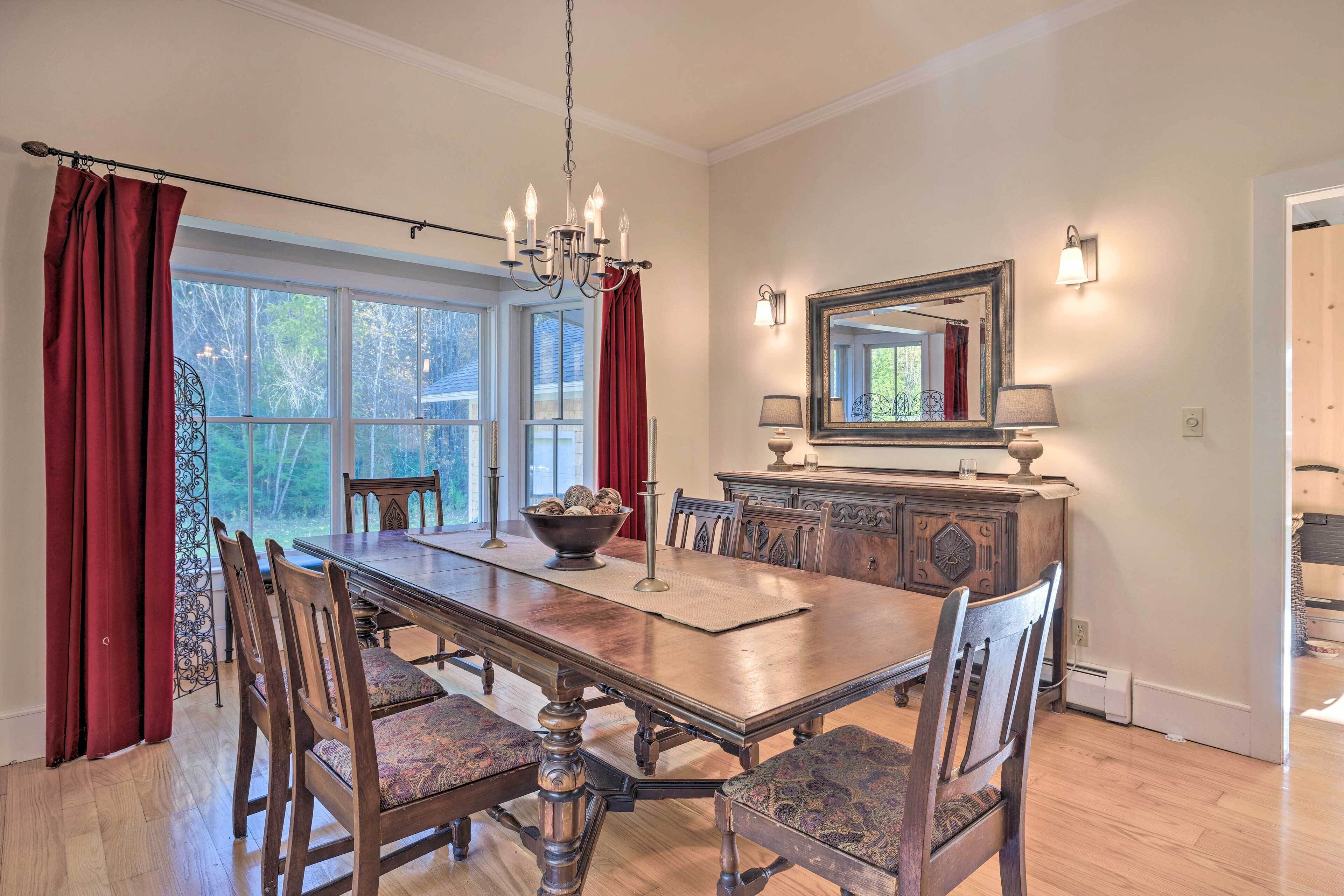 A dining room with a large wooden table, chairs, a chandelier, and a view through large windows.
