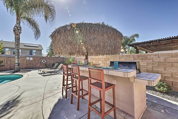 An outdoor bar area with a concrete floor, wooden bar stools, and a pool in the background.