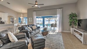 Ein geräumiges Wohnzimmer mit einer Ledersofa, einem Hocker und einem Couchtisch. Ein Fernseher steht auf einem Holzgestell, ein Deckenventilator sorgt für Luftzirkulation und ein großes Fenster ist mit Vorhängen versehen.