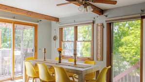 Un coin repas avec une table en bois et des chaises jaunes, de grandes fenêtres donnant sur la verdure et un ventilateur de plafond.