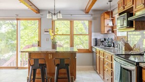 Une cuisine dotée d’un grand îlot central, de meubles en bois et offrant une vue sur les arbres grâce à de grandes fenêtres.