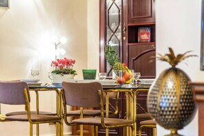 Un coin repas avec une table dressée pour quatre personnes, agrémenté de chaises dorées, d’un vase décoratif et d’un panier de fruits.