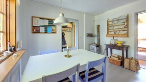 Een eethoek met een witte tafel en stoelen, een hanglamp, een prikbord met papieren en een plank met servies.