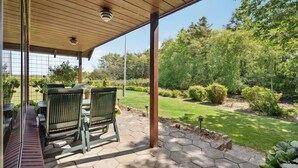 A covered patio with two green chairs and a table, overlooking a well-maintained garden with a lawn and various shrubs. - Nice home in Bækmarksbro with WiFi (Bækmarksbro)