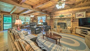 A cozy log cabin interior with a fireplace, leather chairs, a sofa, and a television. - Scenic Creekside Cabin w/ Wraparound Porch! (Highlands)
