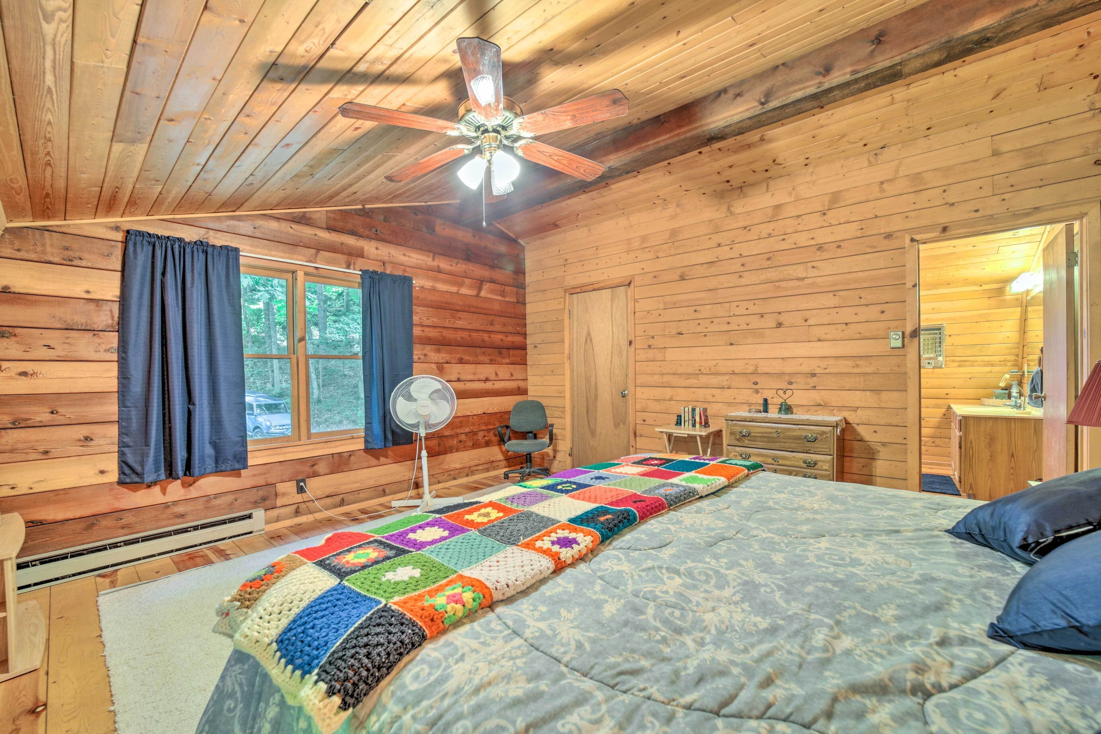 Un dormitorio de cabaña de madera con una cama, un ventilador de techo, una ventana con cortinas, un escritorio y un aparador.