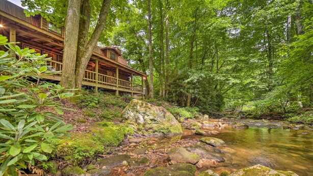 A cabin with a deck overlooking a forested area and a stream. - Scenic Creekside Cabin w/ Wraparound Porch! (Highlands)