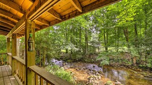 A wooden deck overlooking a forested area with a stream. - Scenic Creekside Cabin w/ Wraparound Porch! (Highlands)