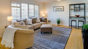 A living room with a beige sofa, a white throw blanket, a striped rug, a wooden floor, a side table with a lamp, a potted plant, and a wall-mounted shelf with glassware. - Downtown Kirkland Rental: Steps to Lake Washington (Kirkland)