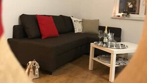 Een moderne woonkamer met een donkere bank, een rood kussen, een witte salontafel met glazen en een fles, en een ingelijst kunstwerk aan de muur.