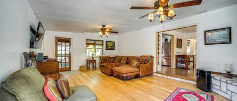 A spacious living room with hardwood flooring, a large sofa, a smaller armchair, and a ceiling fan.