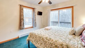 Ein Schlafzimmer mit einem Bett, einem Deckenventilator, zwei Fenstern, einem Fernseher und Blick auf eine Holzterrasse.