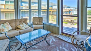 A sunlit room with a beige sofa, a glass coffee table, and a view of a wooden deck and buildings. - Spacious North Topsail Family Home With 2 Decks (Sneads Ferry)