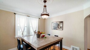 Een eetgedeelte met een houten tafel, zwarte stoelen, een kroonluchter, een raam met transparante gordijnen en een aan de muur hangend schilderij.