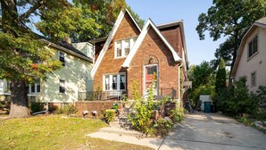 A two-story brick house with a front porch, a small garden, and a driveway leading to a garage. - Ferndale Apartment w/ Yard - Near the Detroit Zoo! (Ferndale)