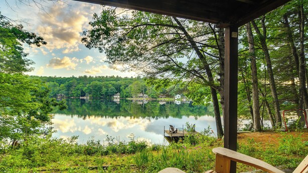 A wooden deck overlooking a calm lake with a forested shoreline and a small dock with a chair. - Waterfront Escape w/ Kayak, Paddle Boards, & Sauna (Casco)