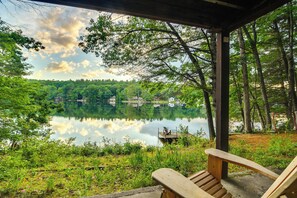 A wooden deck overlooking a calm lake with a forested shoreline and a small dock with a chair.