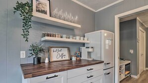 Une cuisine avec des meubles blancs, un plan de travail en bois et une machine à café. On y trouve des étagères avec des bocaux et un tableau encadré au mur.