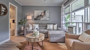 Un salon avec un canapé gris, une table basse ronde et un fauteuil beige. Une grande fenêtre offre une vue sur la verdure à l’extérieur.