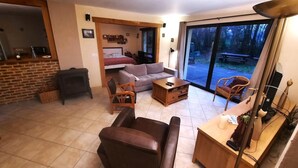 Una sala de estar con sofá, sillón, mesa de centro y chimenea. Se ve una cama al fondo y un ventanal con vistas al exterior.