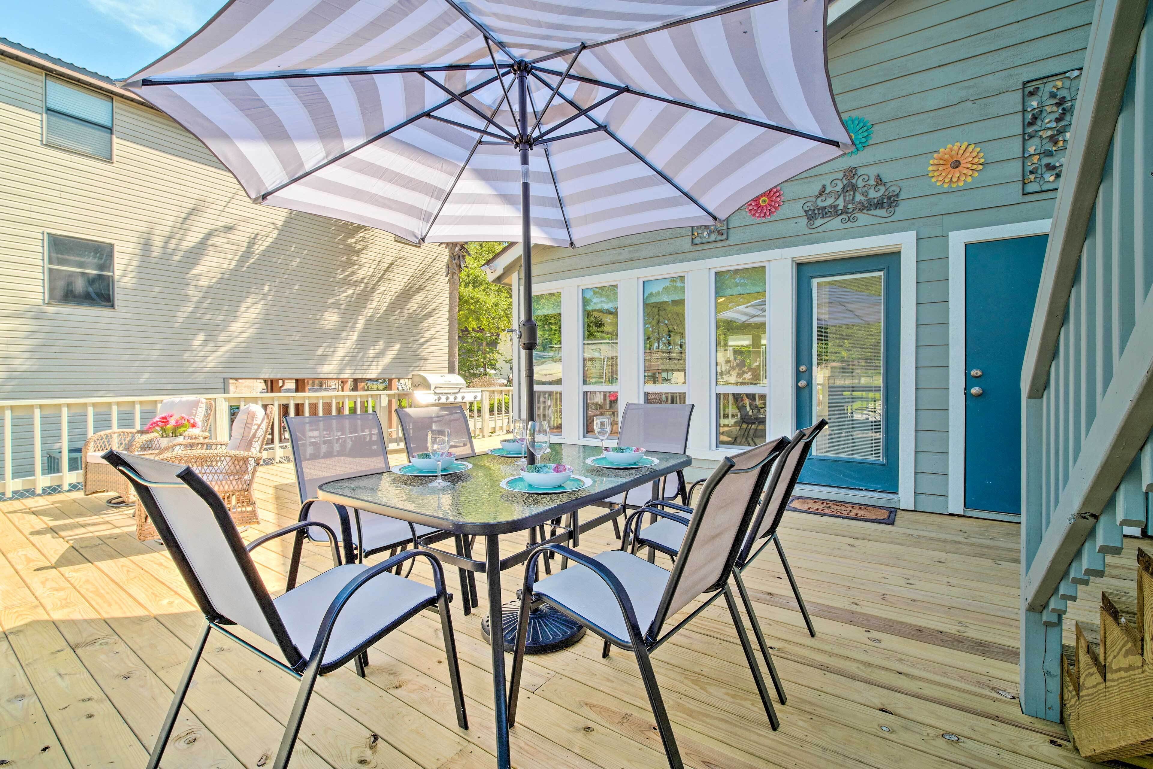 Un espace extérieur abrité, aménagé pour les repas, avec une table et des chaises, un grand parasol et une vue sur une maison avec une porte et des fenêtres bleues.