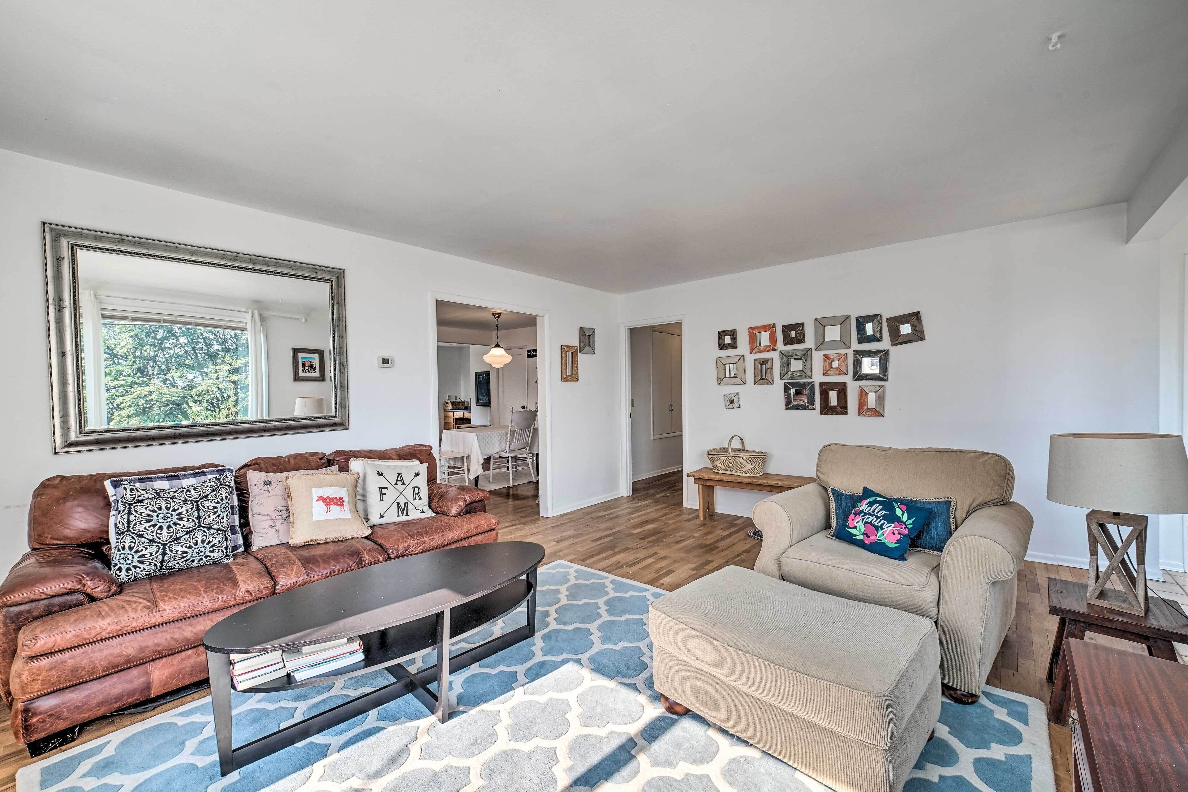 A living room with a brown leather sofa, a beige armchair, a coffee table, and a patterned rug. There are framed pictures on the wall and a lamp on a side table.