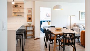 Una cocina moderna con armarios blancos, una mesa de comedor de madera y sillas negras. Hay una lámpara colgante blanca sobre la mesa y una lámpara de techo blanca.