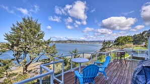 A deck with blue chairs and a table overlooking a lake, with a tree and a distant bridge.