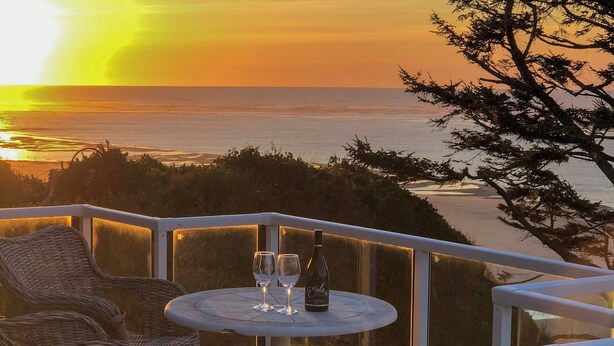 A balcony with wicker chairs and a table, overlooking a sunset over the ocean.