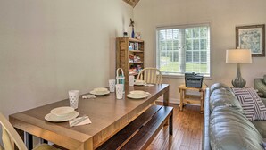 Un comedor con una mesa de madera dispuesta para cuatro personas, una estantería, una ventana con persianas, una lámpara y un sofá con cojines estampados.