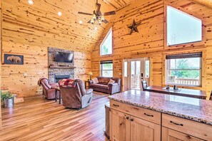 A spacious log cabin interior with a stone fireplace, leather furniture, a kitchen with granite countertops, and large windows offering natural light and views. - Cozy Murphy Cabin Rental w/ Mountain Views! (Murphy)