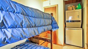 Ein Etagenbett mit blauer Matratze, einer Holzleiter und einer kleinen Holzkassette unter dem Bett. Ein weißer Kühlschrank mit Gefriertruhe steht daneben, und eine Holztür führt in einen anderen Raum.