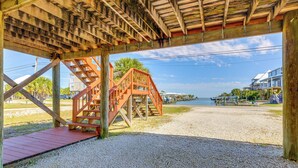 A wooden pier with a staircase leading to a covered area, a gravel path, and a view of a beach and buildings in the background. - Breezy Dauphin Island Vacation Rental With Deck! (Dauphin Island)
