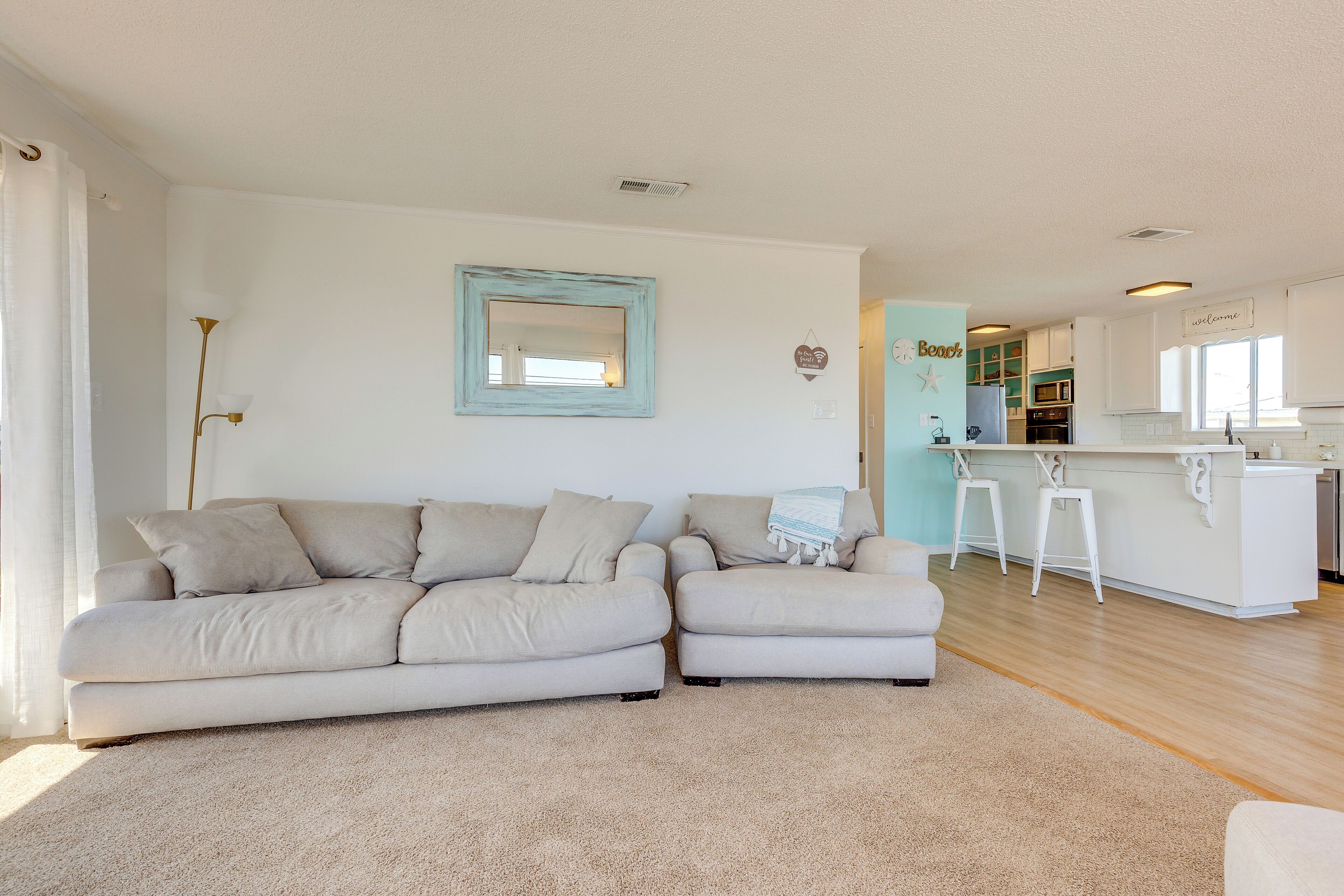 A living room with a beige sectional sofa, a small rug, a wall-mounted mirror, and a kitchen area with white cabinets and a bar stool.