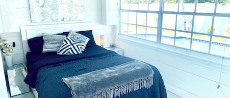 A bedroom with a bed, a nightstand, a window with a view of a pool, and a wall with a mosaic artwork.